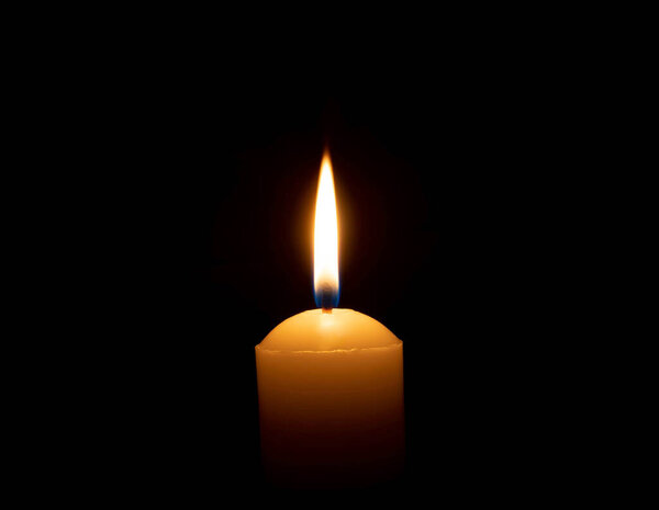 A single burning candle flame or light is glowing on a big yellow candle on black or dark background on table in church for Christmas, funeral or memorial service with copy space