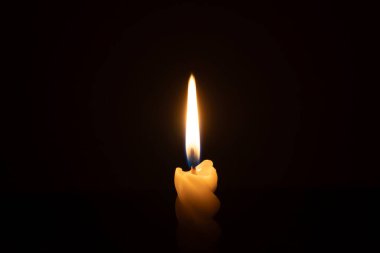 A single burning candle flame or light is glowing on a spiral white candle on black or dark background on table in church for Christmas, funeral or memorial service with copy space clipart