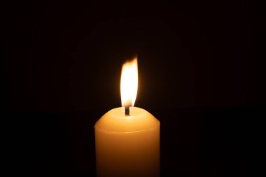 A single burning candle flame or light glowing on a big white candle is isolated on red or dark background on table in church for Christmas, funeral or memorial service clipart