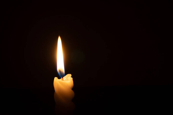 A single burning candle flame or light is glowing on a spiral white candle on black or dark background on table in church for Christmas, funeral or memorial service with copy space