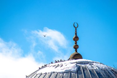 Hilal ay sembollü bir İslam camisinin kubbesi, kar aksanları, güvercinler, mavi gökyüzüne karşı tünemiş..