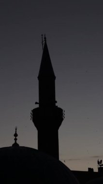 Gün batımında bir caminin silueti, kuşlar uçarken, İslami günler için ideal dikey metin alanı, sükuneti ve ruhani yansımayı sembolize ediyor..