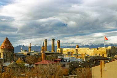 The historic skyline of Erzurum with its iconic castle, Twin Minaret Madrasa, mosques, and tombs, set against a dramatic sky and snowy mountain backdrop.  clipart