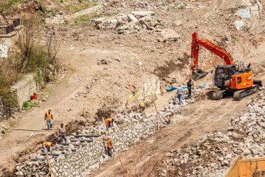 Türkiye 'nin Erzurum kentinde kentsel yenileme çalışmaları sırasında taş duvar inşa eden bir kazıcı işçilere yardım ediyor. Dönüşüm, takım çalışması ve altyapı ilerlemesini sembolize ediyor.