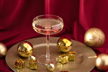 Mutlu Noeller ve mutlu yıllar. Kırmızı kadife perdenin üzerinde altın Noel süsleri olan bir kadeh şampanya..