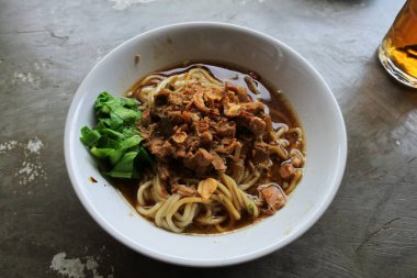 Beyaz kasede tavuklu erişte ve arka planda ahşap çubuklar tavuk şeridi, sebze, köfte, kırmızı biber sosu ve tuzlu kraker. Mie Ayam Endonezya Eriştesi