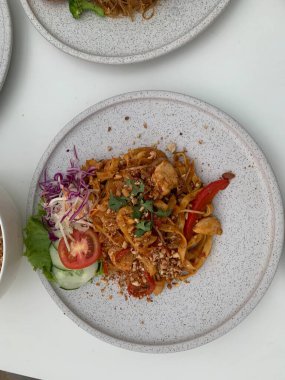 Pişmiş erişteyle kızartılmış beyaz tabak yeşil sebzeler taze marul, salatalık, brokoli ve domates dilimi ile karıştırılmış tavuk, yumurta veya tofu ile süslenmiş. Çatal eriştenin üstüne konur..