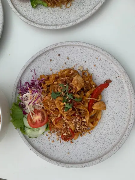 Pişmiş erişteyle kızartılmış beyaz tabak yeşil sebzeler taze marul, salatalık, brokoli ve domates dilimi ile karıştırılmış tavuk, yumurta veya tofu ile süslenmiş. Çatal eriştenin üstüne konur..