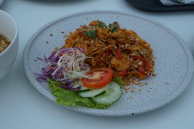 Pişmiş erişteyle kızartılmış beyaz tabak yeşil sebzeler taze marul, salatalık, brokoli ve domates dilimi ile karıştırılmış tavuk, yumurta veya tofu ile süslenmiş. Çatal eriştenin üstüne konur..