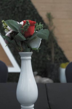 Beyaz bir seramik vazoda çiçek aranjmanı vardır. Koyu yeşil yapraklı kırmızı güller, çeşitli çiçekler siyah masa örtüsü üzerine konur. Modern sanatsal ortam oluşturuluyor.