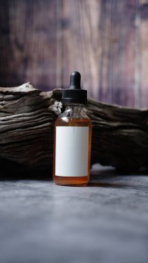 Siyah damperli kapaklı küçük bir cam şişe aşınmış ahşap parçasına dayanır, kehribar renkli bir sıvı içerir ve boş beyaz etiket içerir..