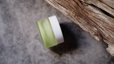 Açık yeşil sıvı madde içeren küçük cam kavanoz, aşınmış ahşap parçasına dayanır ve bileşime dengeli estetik yaratan doğal element ekler.. 