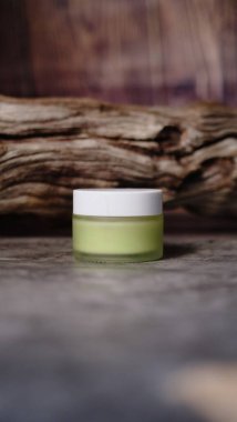 Açık yeşil sıvı madde içeren küçük cam kavanoz, aşınmış ahşap parçasına dayanır ve bileşime dengeli estetik yaratan doğal element ekler.. 