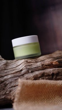 Açık yeşil sıvı madde içeren küçük cam kavanoz, aşınmış ahşap parçasına dayanır ve bileşime dengeli estetik yaratan doğal element ekler.. 