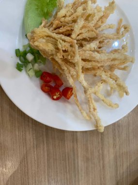 Çıtır çıtır kızarmış enoki mantarları, yanında taze marul dilimlenmiş yeşil soğan ve doğranmış kırmızı biber ile birlikte beyaz tabakta düzenlenir.. 