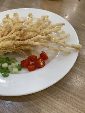 Çıtır çıtır kızarmış enoki mantarları, yanında taze marul dilimlenmiş yeşil soğan ve doğranmış kırmızı biber ile birlikte beyaz tabakta düzenlenir.. 