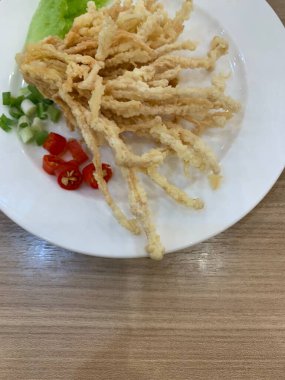 Çıtır çıtır kızarmış enoki mantarları, yanında taze marul dilimlenmiş yeşil soğan ve doğranmış kırmızı biber ile birlikte beyaz tabakta düzenlenir.. 