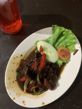 Siyah biberli pişirilmiş et oval beyaz tabakta servis edilir koyu ahşap ve yanında taze marul ve salatalık dilimi. Tabakta dilimlenmiş kırmızı biber ve soğan var..