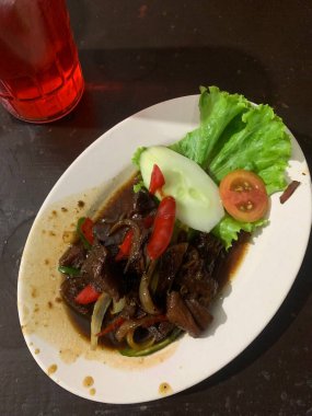 Siyah biberli pişirilmiş et oval beyaz tabakta servis edilir koyu ahşap ve yanında taze marul ve salatalık dilimi. Tabakta dilimlenmiş kırmızı biber ve soğan var..