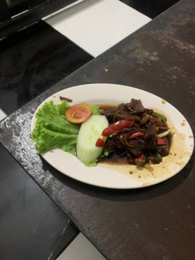 Siyah biberli pişirilmiş et oval beyaz tabakta servis edilir koyu ahşap ve yanında taze marul ve salatalık dilimi. Tabakta dilimlenmiş kırmızı biber ve soğan var..