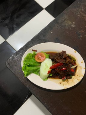 Siyah biberli pişirilmiş et oval beyaz tabakta servis edilir koyu ahşap ve yanında taze marul ve salatalık dilimi. Tabakta dilimlenmiş kırmızı biber ve soğan var..