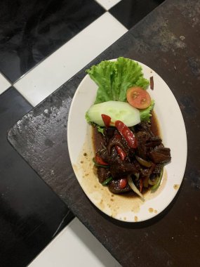 Siyah biberli pişirilmiş et oval beyaz tabakta servis edilir koyu ahşap ve yanında taze marul ve salatalık dilimi. Tabakta dilimlenmiş kırmızı biber ve soğan var..