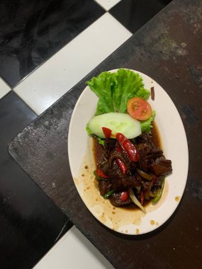 Siyah biberli pişirilmiş et oval beyaz tabakta servis edilir koyu ahşap ve yanında taze marul ve salatalık dilimi. Tabakta dilimlenmiş kırmızı biber ve soğan var..