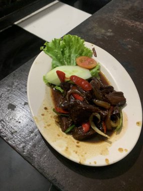 Siyah biberli pişirilmiş et oval beyaz tabakta servis edilir koyu ahşap ve yanında taze marul ve salatalık dilimi. Tabakta dilimlenmiş kırmızı biber ve soğan var..