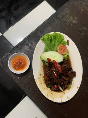 Siyah biberli pişirilmiş et oval beyaz tabakta servis edilir koyu ahşap ve yanında taze marul ve salatalık dilimi. Tabakta dilimlenmiş kırmızı biber ve soğan var..