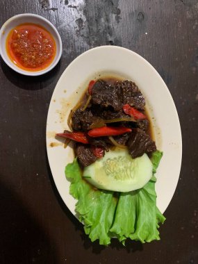 Siyah biberli pişirilmiş et oval beyaz tabakta servis edilir koyu ahşap ve yanında taze marul ve salatalık dilimi. Tabakta dilimlenmiş kırmızı biber ve soğan var..