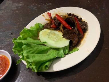 Siyah biberli pişirilmiş et oval beyaz tabakta servis edilir koyu ahşap ve yanında taze marul ve salatalık dilimi. Tabakta dilimlenmiş kırmızı biber ve soğan var..