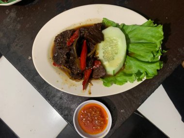 Siyah biberli pişirilmiş et oval beyaz tabakta servis edilir koyu ahşap ve yanında taze marul ve salatalık dilimi. Tabakta dilimlenmiş kırmızı biber ve soğan var..