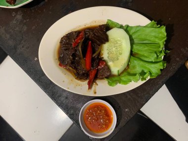 Siyah biberli pişirilmiş et oval beyaz tabakta servis edilir koyu ahşap ve yanında taze marul ve salatalık dilimi. Tabakta dilimlenmiş kırmızı biber ve soğan var..