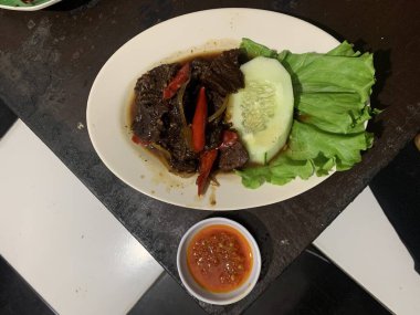 Siyah biberli pişirilmiş et oval beyaz tabakta servis edilir koyu ahşap ve yanında taze marul ve salatalık dilimi. Tabakta dilimlenmiş kırmızı biber ve soğan var..