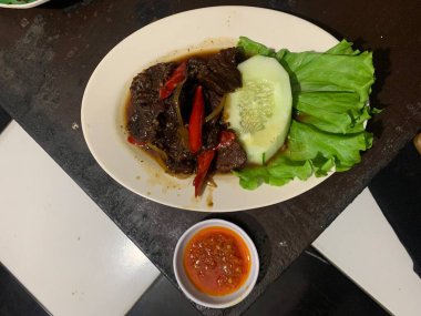 Siyah biberli pişirilmiş et oval beyaz tabakta servis edilir koyu ahşap ve yanında taze marul ve salatalık dilimi. Tabakta dilimlenmiş kırmızı biber ve soğan var..