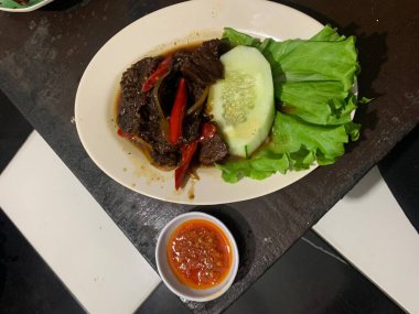 Siyah biberli pişirilmiş et oval beyaz tabakta servis edilir koyu ahşap ve yanında taze marul ve salatalık dilimi. Tabakta dilimlenmiş kırmızı biber ve soğan var..