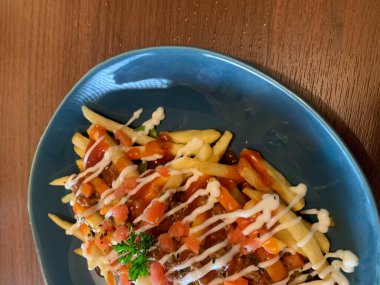 Kızarmış patatesler çeşitli soslarla süslenmiş, domates ve taze maydanozla süslenmiş. Ahşap masaya konmuş gri tabakta servis edilir..