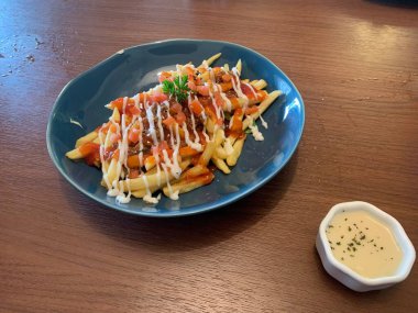 Kızarmış patatesler çeşitli soslarla süslenmiş, domates ve taze maydanozla süslenmiş. Ahşap masaya konmuş gri tabakta servis edilir..