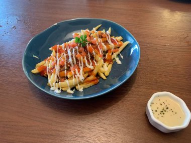 Kızarmış patatesler çeşitli soslarla süslenmiş, domates ve taze maydanozla süslenmiş. Ahşap masaya konmuş gri tabakta servis edilir..