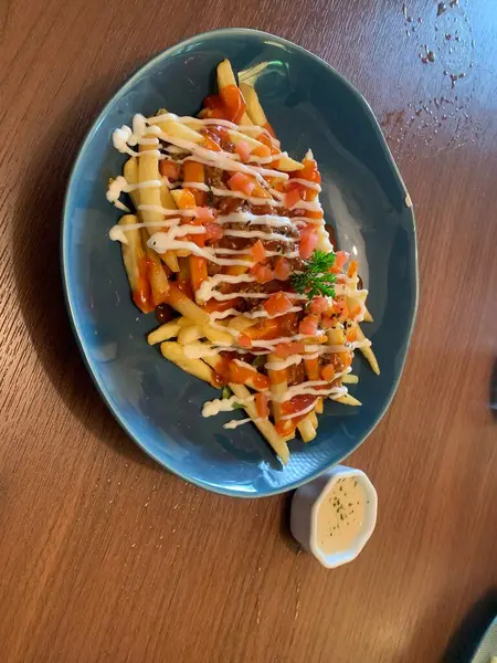 Kızarmış patatesler çeşitli soslarla süslenmiş, domates ve taze maydanozla süslenmiş. Ahşap masaya konmuş gri tabakta servis edilir..