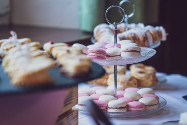 Cam pasta standına yerleştirilmiş harika pembe ve beyaz badem kurabiyelerinin hoş bir karışımı yumuşak odaklı arka planda diğer hamur işleriyle tamamlanmış büyüleyici ve zarif bir görsel cazibe yaratıyor..