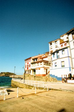 Açık mavi gökyüzünün altında, deniz kenarında renkli binaları olan bir sahil kasabası..