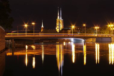 A nighttime scene featuring a tall bridge with lights, a large church illuminated by streetlights, and a tranquil river reflecting the lights. clipart