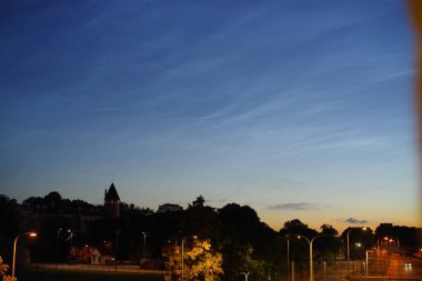 Yıldızlar ve bulutlarla dolu bir gece gökyüzü, uzakta yanan binalar ve koyu mavi gökyüzü olan bir şehir silueti..