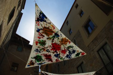 Üzerinde renkli resimler olan yelkenli şekilli bir bayrak binaların arasında asılı, açık bir gökyüzüne karşı..