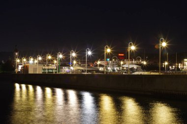 Gece vakti bir liman manzarası, sokak lambaları suya yansıyor ve bir rıhtımı aydınlatıyor..