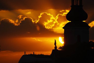 Büyüleyici bir günbatımı şehir manzarasına dramatik gölgeler düşürüyor. Önde gelen bir kilise kulesi ve renkli bulutlar var.