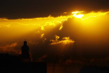 Güneşin kısmen bulutlarla karardığı renkli bir gün batımı binaların ufuk çizgisine ve kilise kulesine vuruyor..
