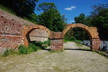 Arkasında çimenlik bir alan olan taştan bir kemer yolu, eski bir tuğla binaya açılıyor..
