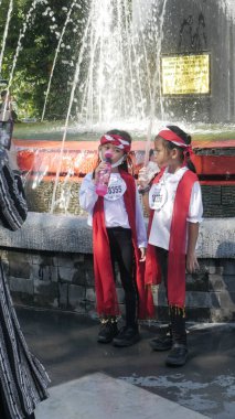 Kırmızı şallı iki çocuk çeşmenin önünde su içiyor. Surabaya, Doğu Java, Endonezya. 18 Aralık 2022